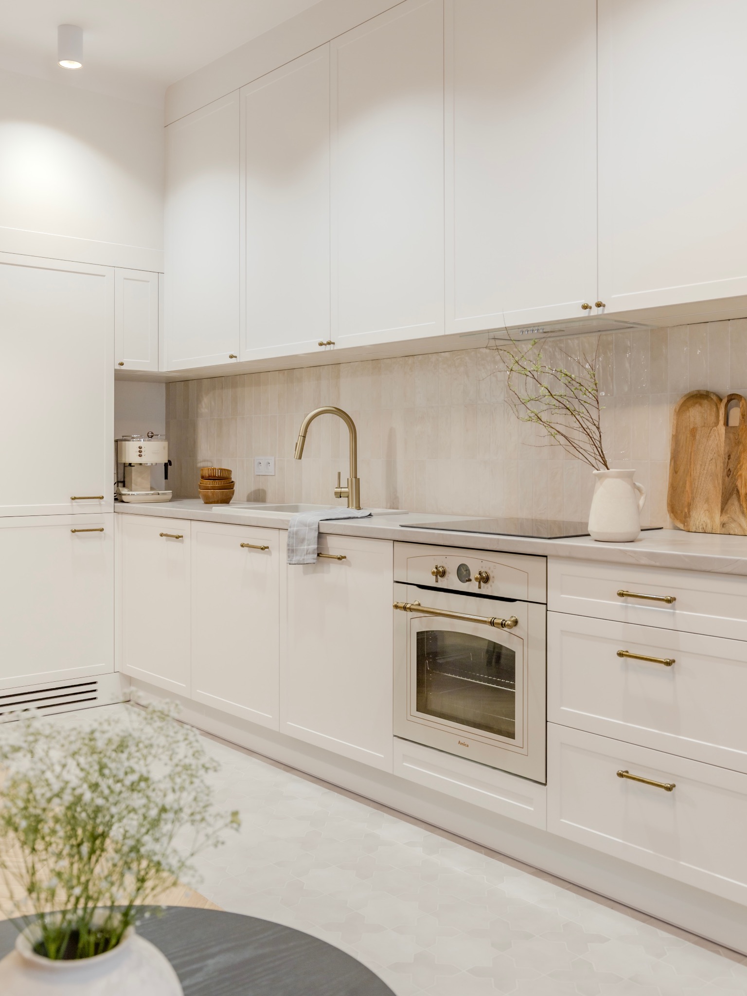 Jasna kuchnia z białymi szafkami, złotą armaturą i sprzętami AGD w stylu retro, z blatem wykończonym w jasnym odcieniu i dekoracyjnymi elementami takimi jak drewniane deski do krojenia i gałązki...