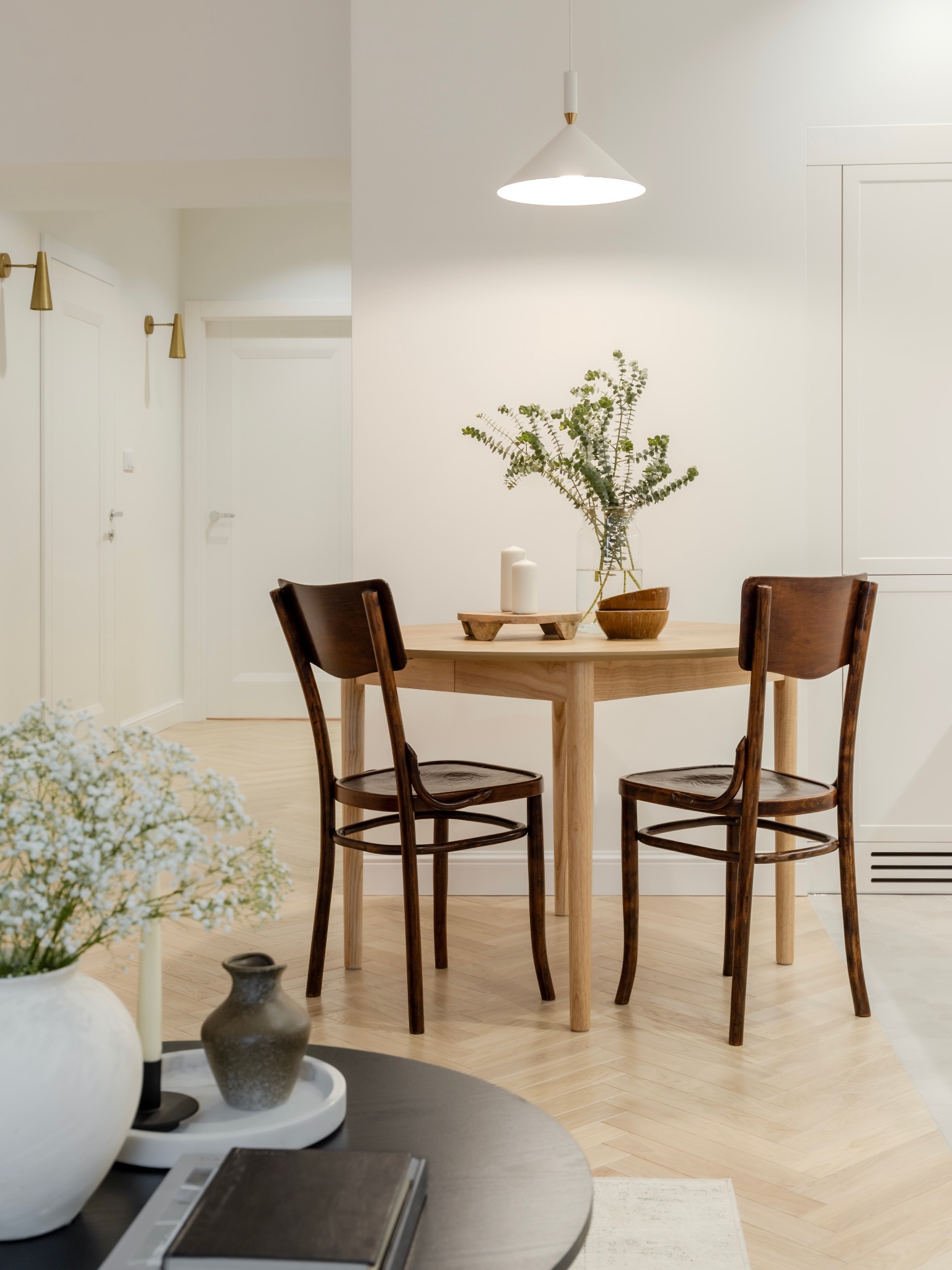 Jasne wnętrze jadalni z okrągłym stołem z drewna, dwoma krzesłami w stylu vintage, minimalistyczną lampą wiszącą i parkietem ułożonym w jodełkę.