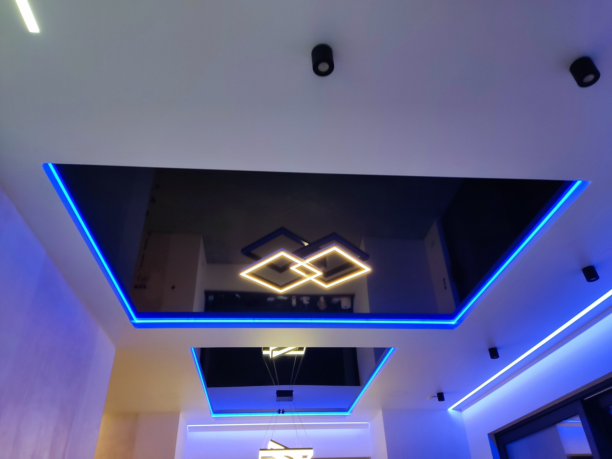 Sufit podwieszany z czarnym lustrem, oświetleniem LED w kolorze niebieskim i nowoczesną lampą w kształcie połączonych kwadratów, widziany z dołu.