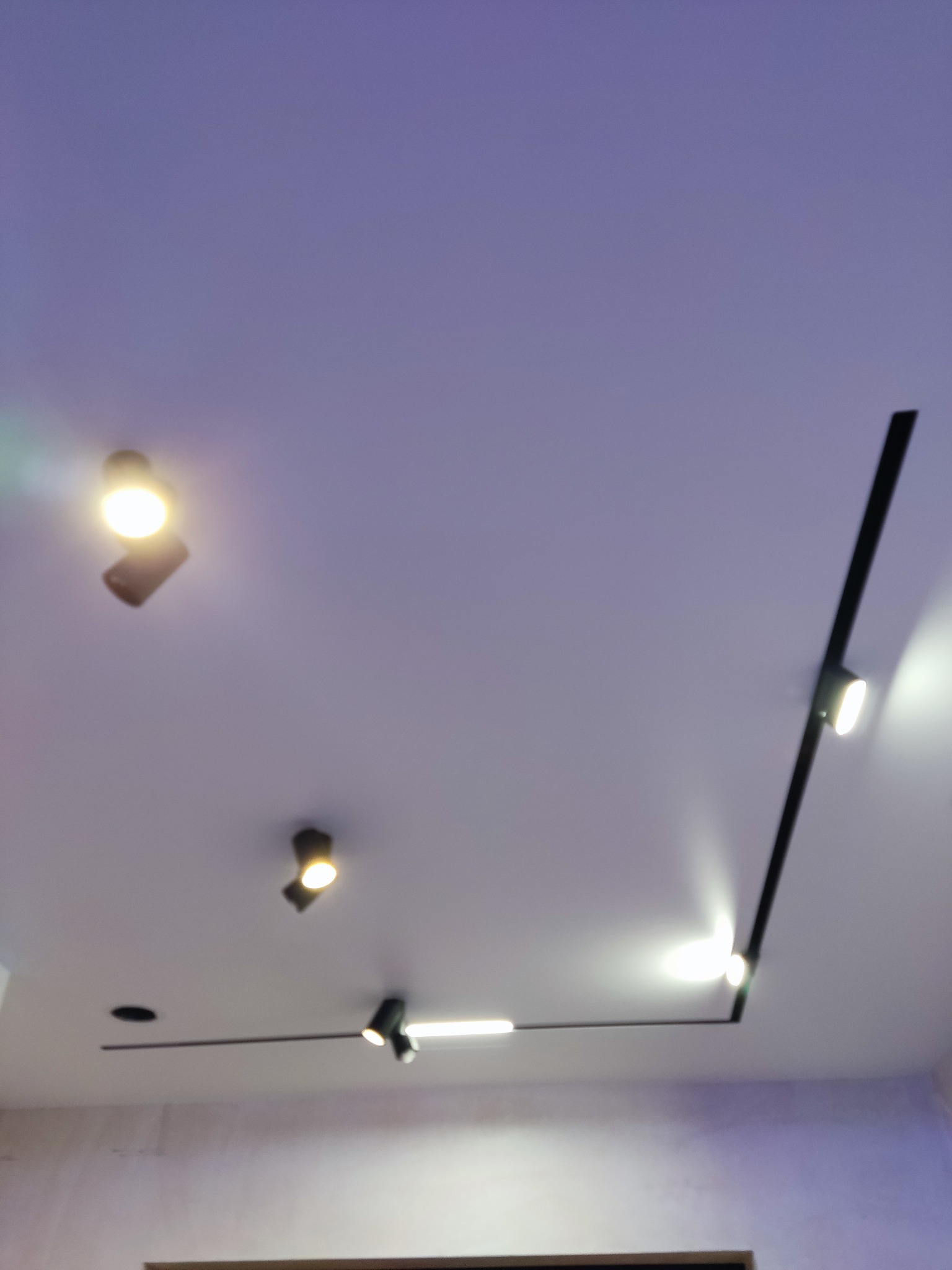 Biały sufit z zamontowanym systemem szynowym oświetleniowym z czarnymi reflektorami punktowymi i liniowymi LED, skierowanymi w dół, włączone światło