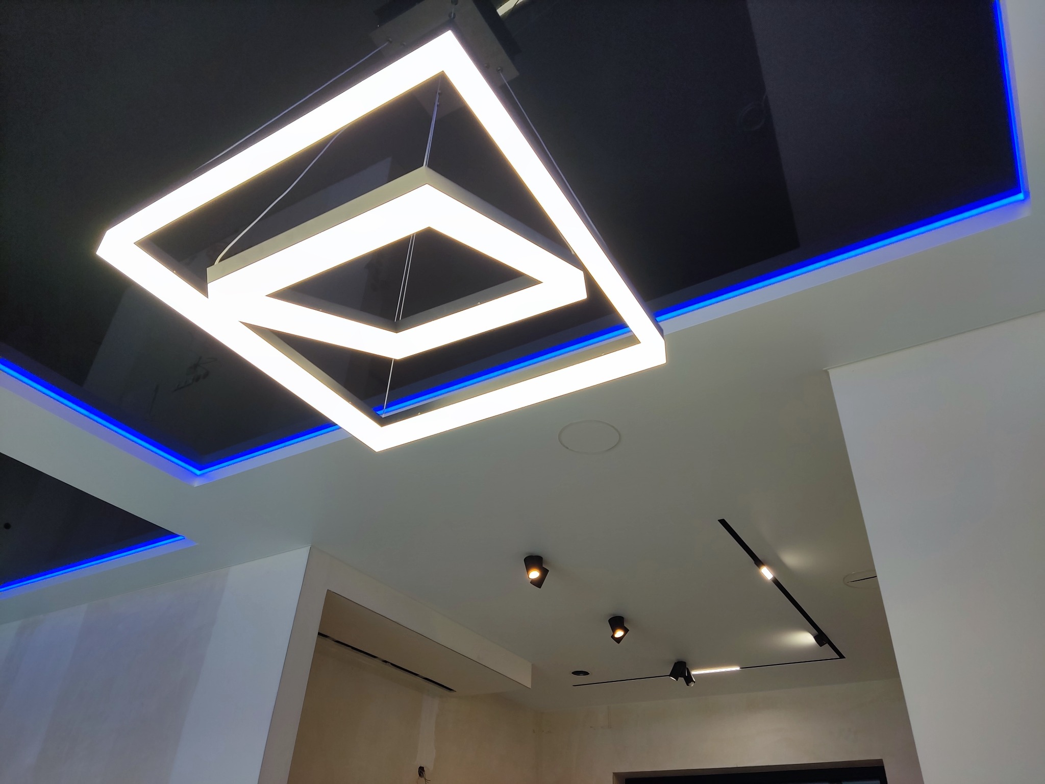 Nowoczesny sufit podwieszany z czarną błyszczącą powierzchnią, oświetleniem LED w kolorze niebieskim i geometryczną lampą wiszącą w kształcie kwadratu z białym światłem, uzupełniony reflektorami...