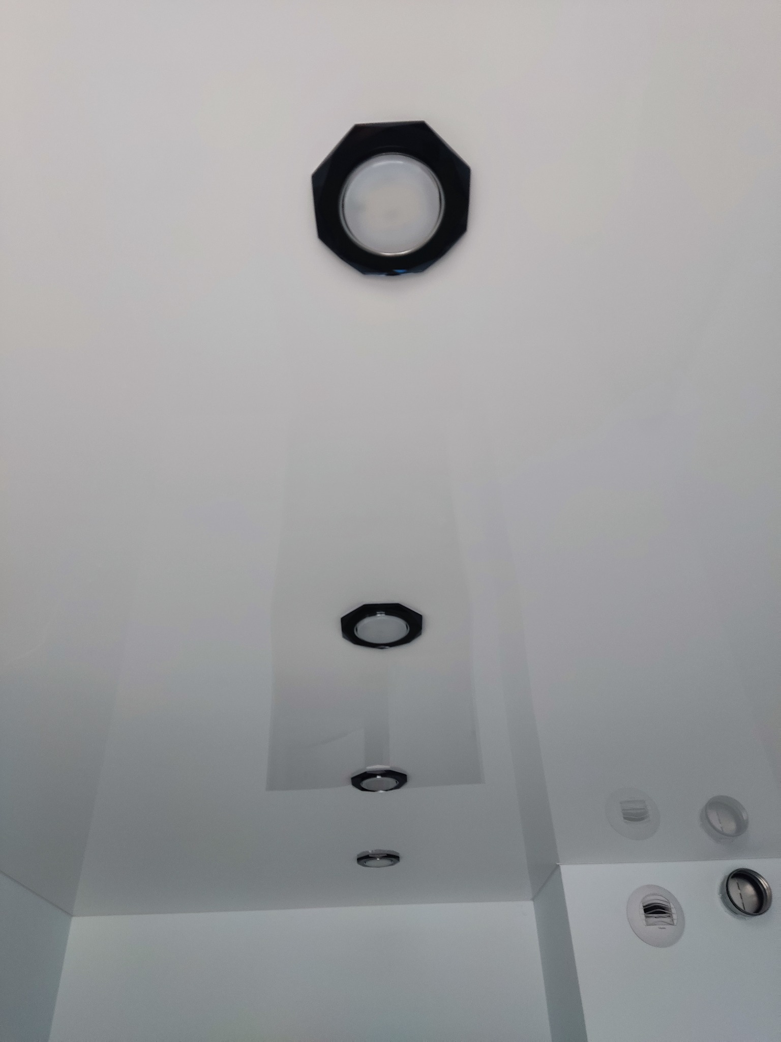 Biały sufit z zamontowanymi, okrągłymi lampami w czarnych, ośmiokątnych obudowach, dodatkowo widoczne okrągłe otwory wentylacyjne.