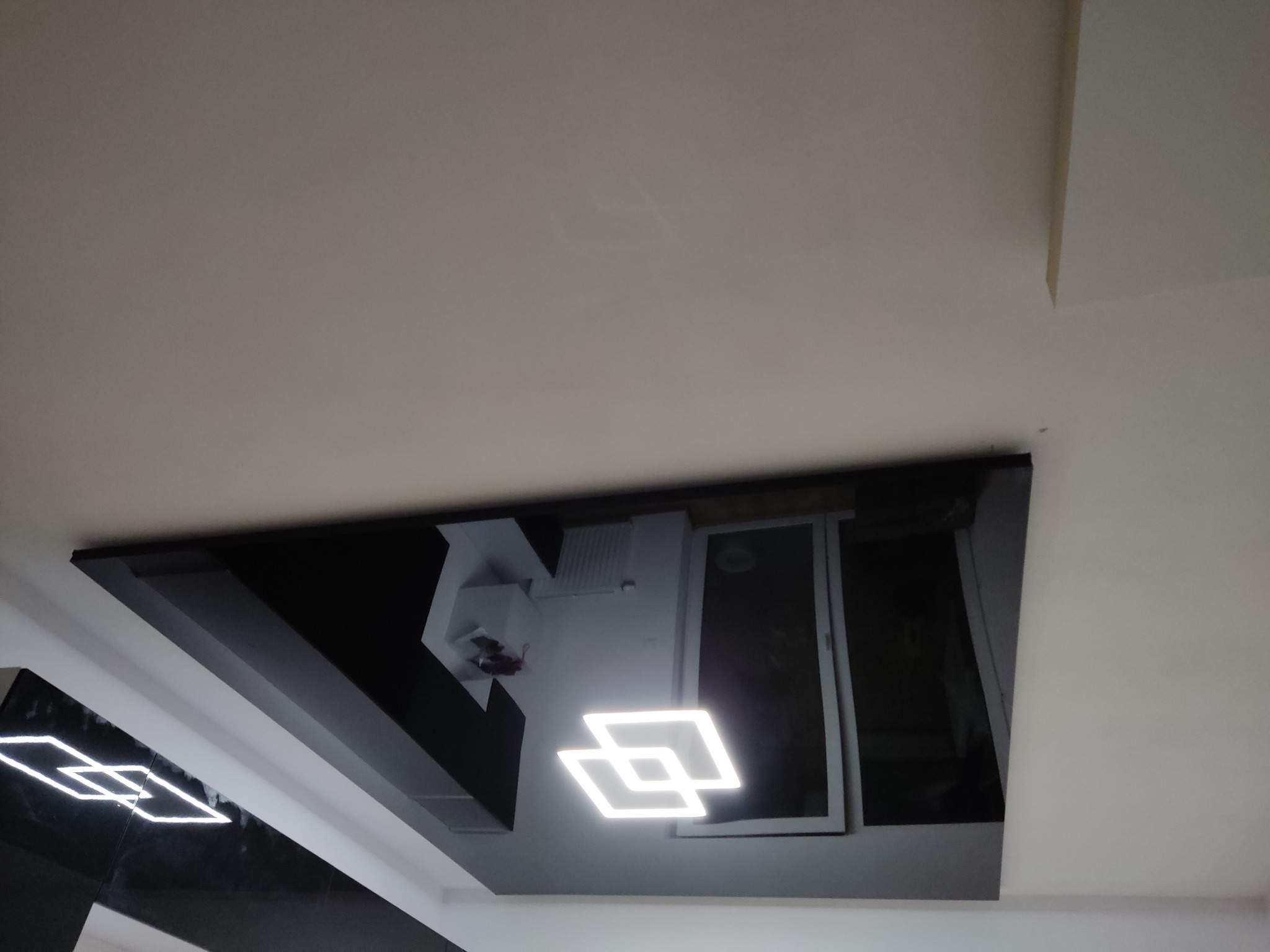 Czarny, podwieszany sufit z błyszczącą powierzchnią i nowoczesnym oświetleniem LED w kształcie kwadratów, odbijający elementy wyposażenia wnętrza.