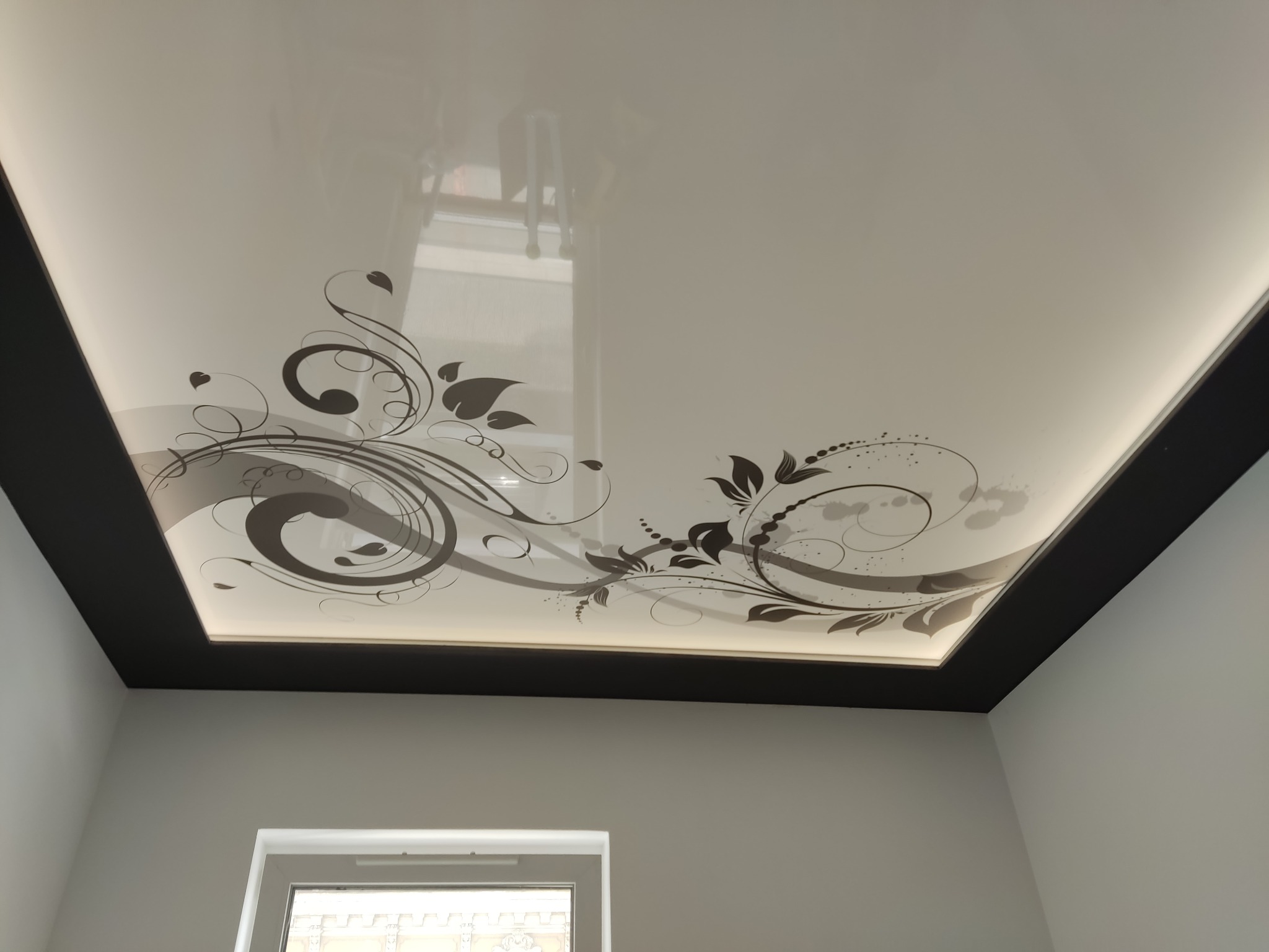 Biały sufit napinany z dekoracyjnym motywem roślinnym w odcieniach szarości, obramowany czarną listwą, widoczne odbicie drabiny.