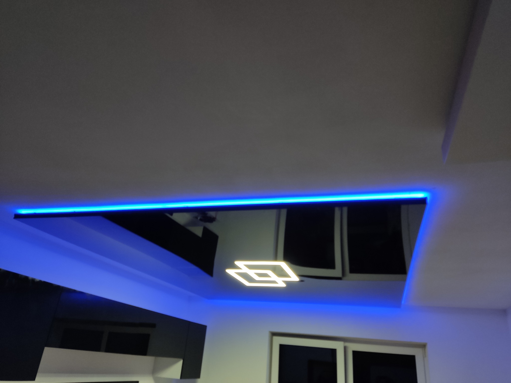 Podwieszany sufit z czarną lustrzaną powierzchnią, oświetlony niebieskim paskiem LED i nowoczesną lampą w kształcie dwóch rombów