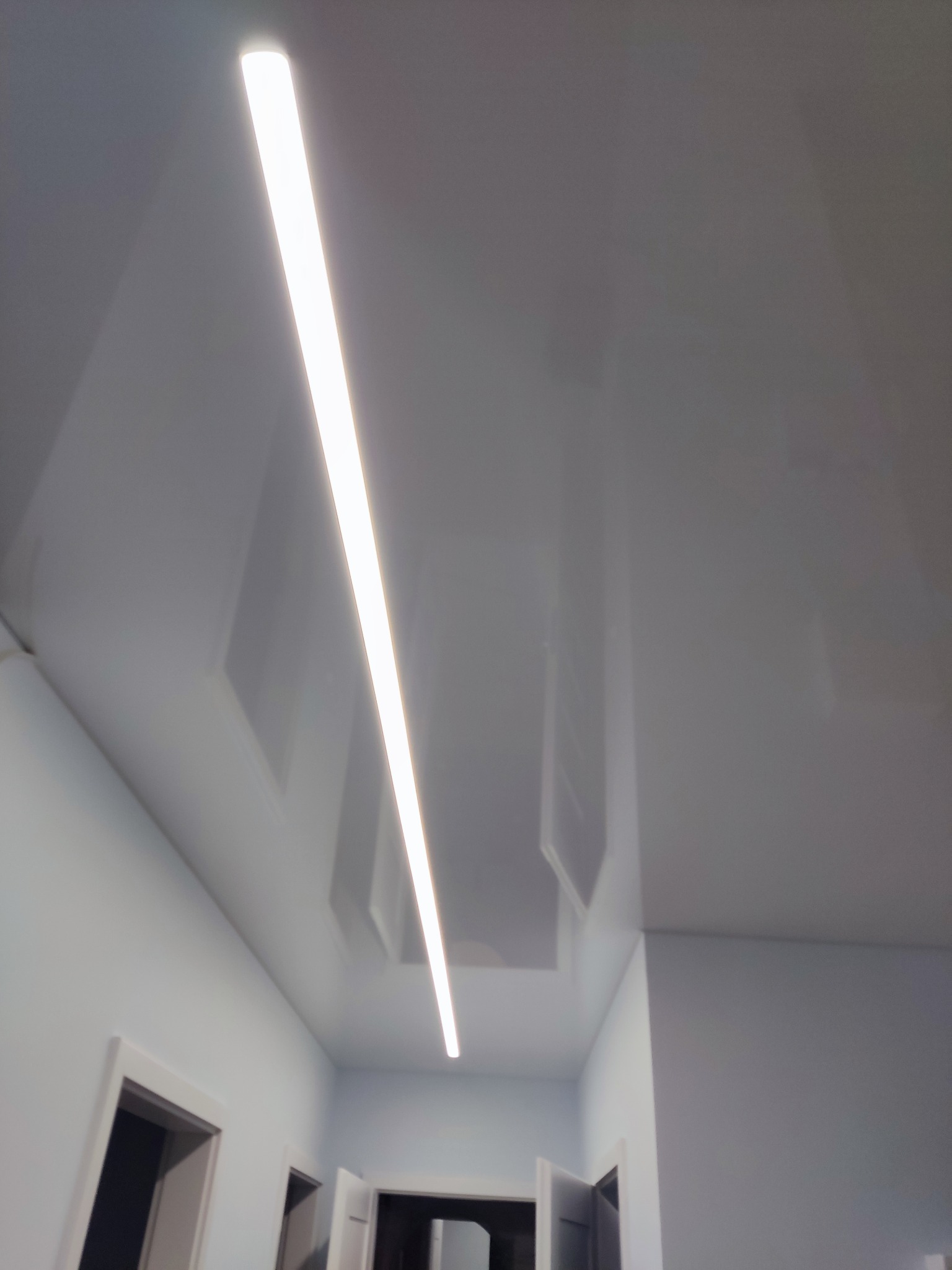 Biały, błyszczący sufit napinany z wbudowanym, liniowym oświetleniem LED, widok z dołu na korytarz z otwartymi drzwiami.