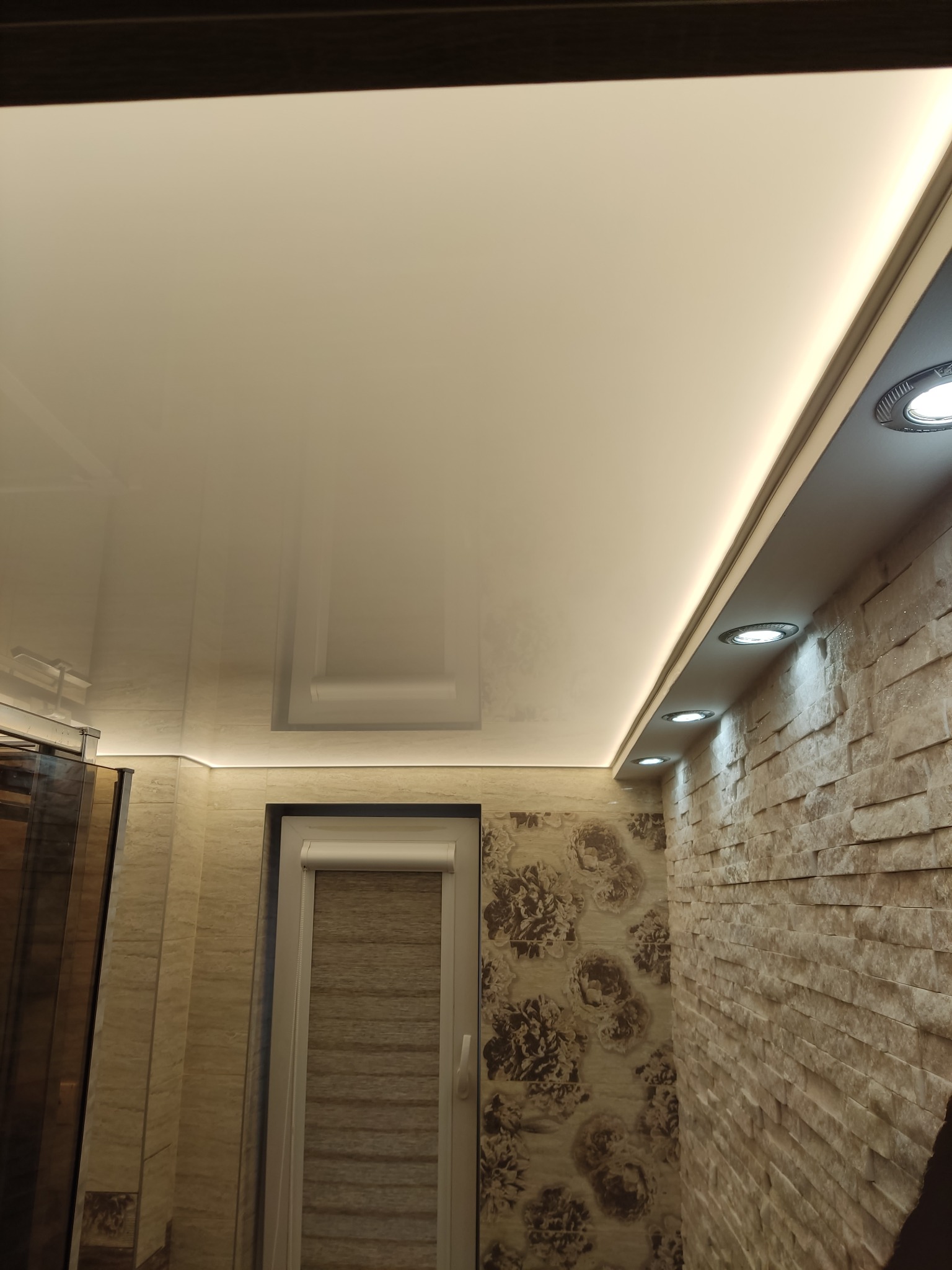 Błyszczący, kremowy sufit napinany z wbudowanym oświetleniem LED, widok z dołu na fragment ściany z kamienia dekoracyjnego i okno z roletą.