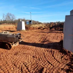 Teren budowy z ubijarką wibro płytową, przygotowany pod wylanie fundamentów, widoczne betonowe podpory i wygładzona powierzchnia ziemi. W tle drzewa i niebieskie niebo.