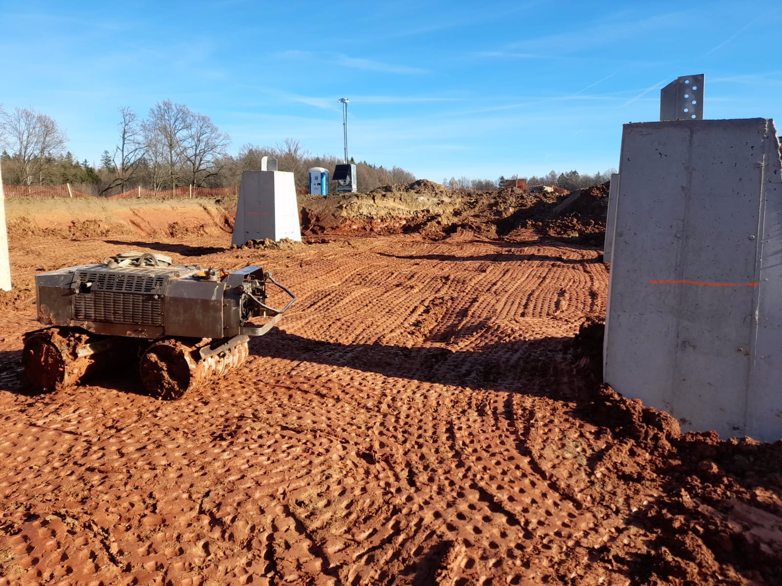 Teren budowy z ubijarką wibro płytową, przygotowany pod wylanie fundamentów, widoczne betonowe podpory i wygładzona powierzchnia ziemi. W tle drzewa i niebieskie niebo.
