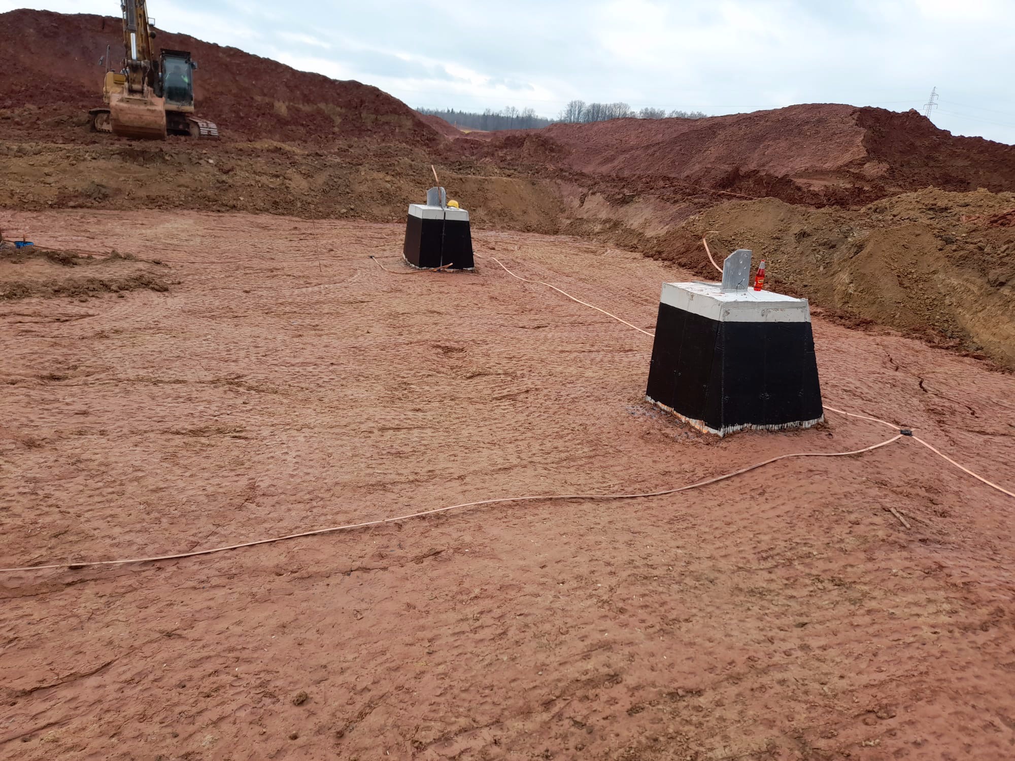 Przygotowany teren pod budowę z widocznymi betonowymi słupami, wykopem ziemnym i koparką w tle.