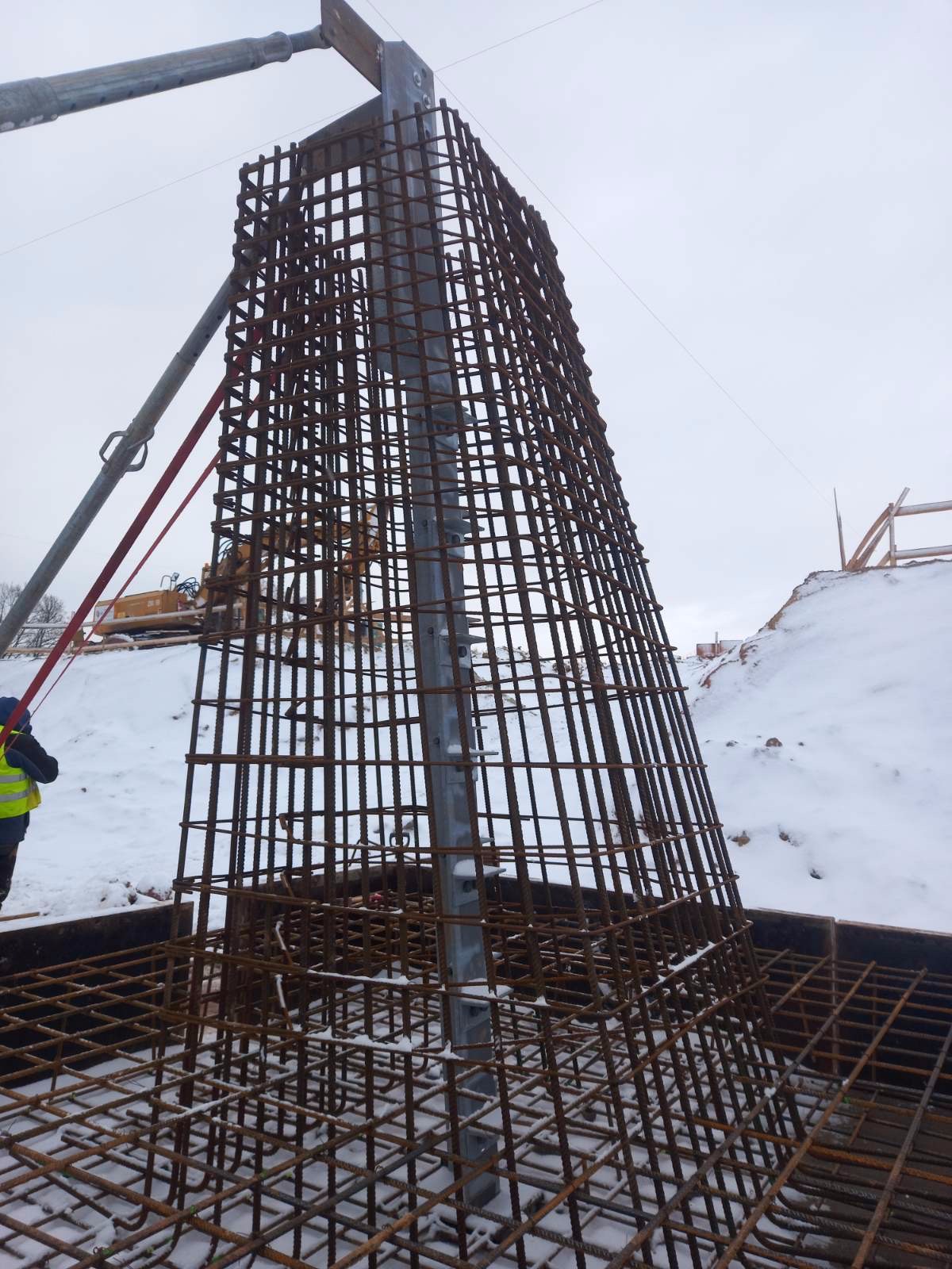 Zimowy krajobraz budowy w Kościanie: stalowe zbrojenie fundamentu w kształcie ściętego ostrosłupa, przygotowane do zalania betonem, z widocznym metalowym słupem wewnątrz i śniegiem w tle.