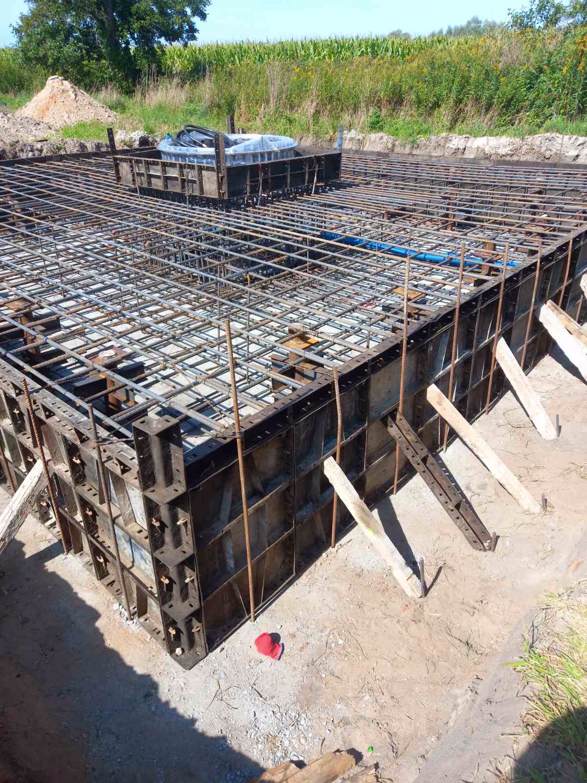 Zbrojenie i szalunek pod płytę fundamentową domu jednorodzinnego z widoczną niebieską rurą instalacyjną, wsparte drewnianymi stemplami, na tle pola uprawnego.