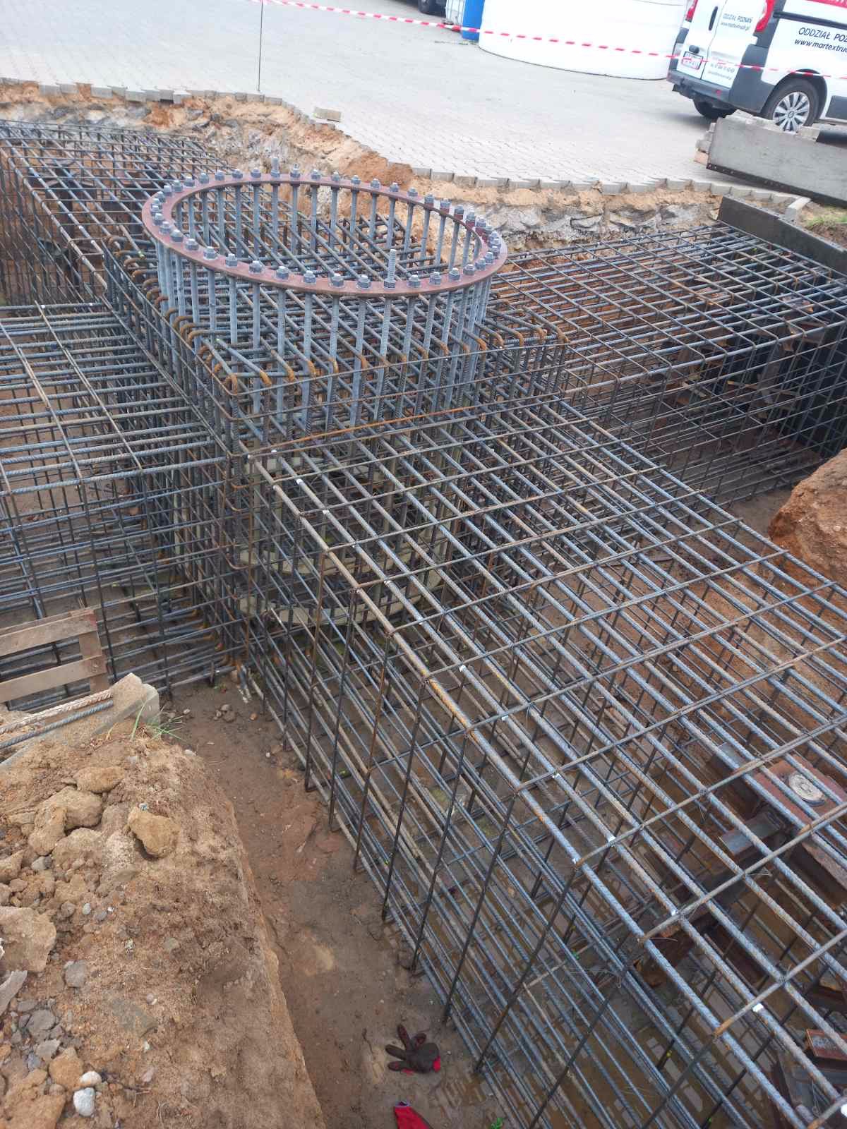 Siatka zbrojeniowa fundamentów z okrągłym elementem wzmacniającym, widoczne wykopane ściany i częściowo utwardzony teren w tle.