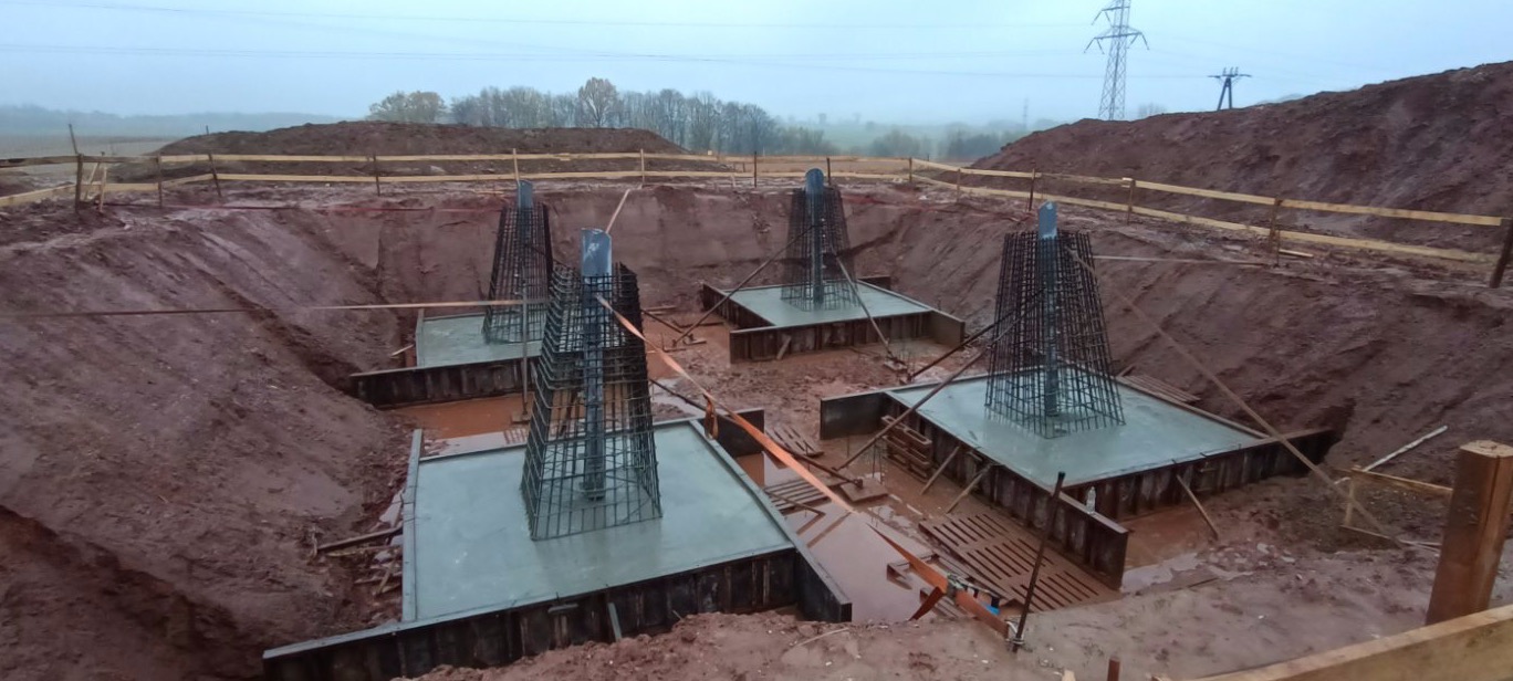 Wykop z czterema betonowymi fundamentami pod budowę, zbrojenie w kształcie stożka, drewniane szalunki i ziemia wokół, pochmurna pogoda.