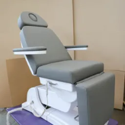 Szare, skórzane krzesło kosmetyczne z regulacją elektryczną, widoczne białe elementy konstrukcyjne i pilot sterujący na fioletowym podłożu, tło z kartonów.