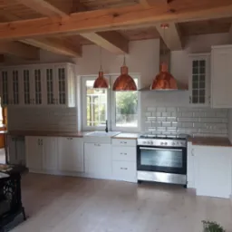 Jasna kuchnia w stylu rustykalnym z drewnianym stropem, białymi szafkami i miedzianymi lampami wiszącymi nad kuchenką.