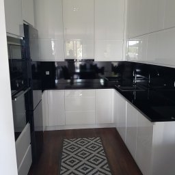 Salon Meblowy Korona - Biała, nowoczesna kuchnia z błyszczącymi frontami szafek, czarnym blatem i panelem nad blatem, oświetleniem punktowym i ciemną podłogą z dywanikiem w geometryczne wzory.