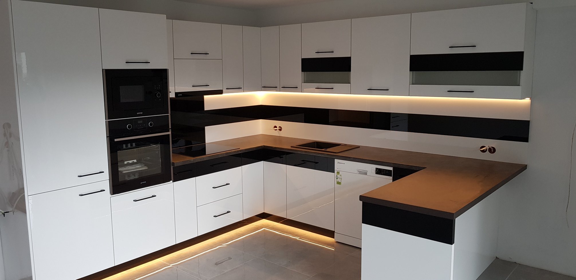 Nowoczesna kuchnia z białymi, lakierowanymi frontami, czarnymi akcentami i oświetleniem LED pod szafkami i na podłodze, z wbudowanym sprzętem AGD marki Gorenje.