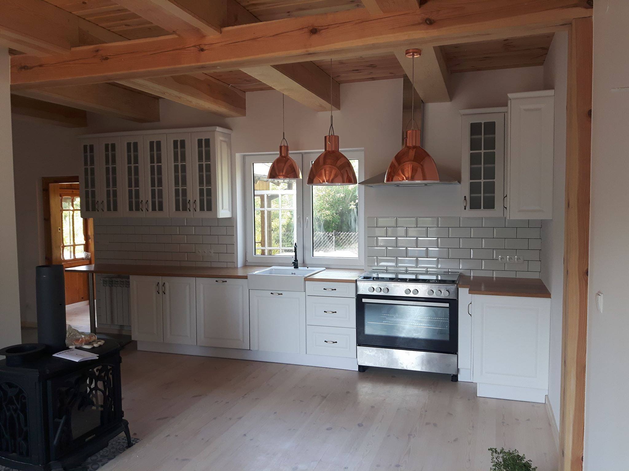 Jasna kuchnia w stylu rustykalnym z drewnianym stropem, białymi szafkami i miedzianymi lampami wiszącymi nad kuchenką.