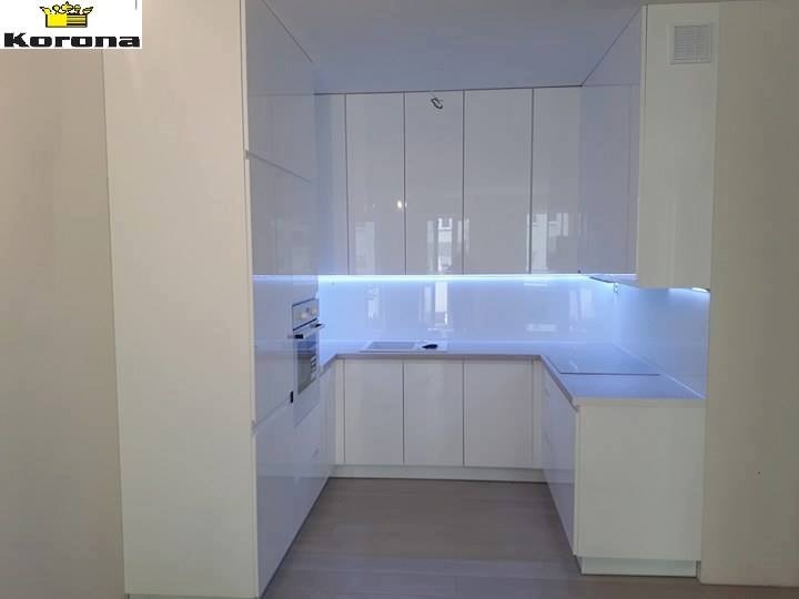 Biała, nowoczesna kuchnia z połyskiem, podświetlana listwą LED pod szafkami wiszącymi, widoczne logo firmy w lewym górnym rogu.