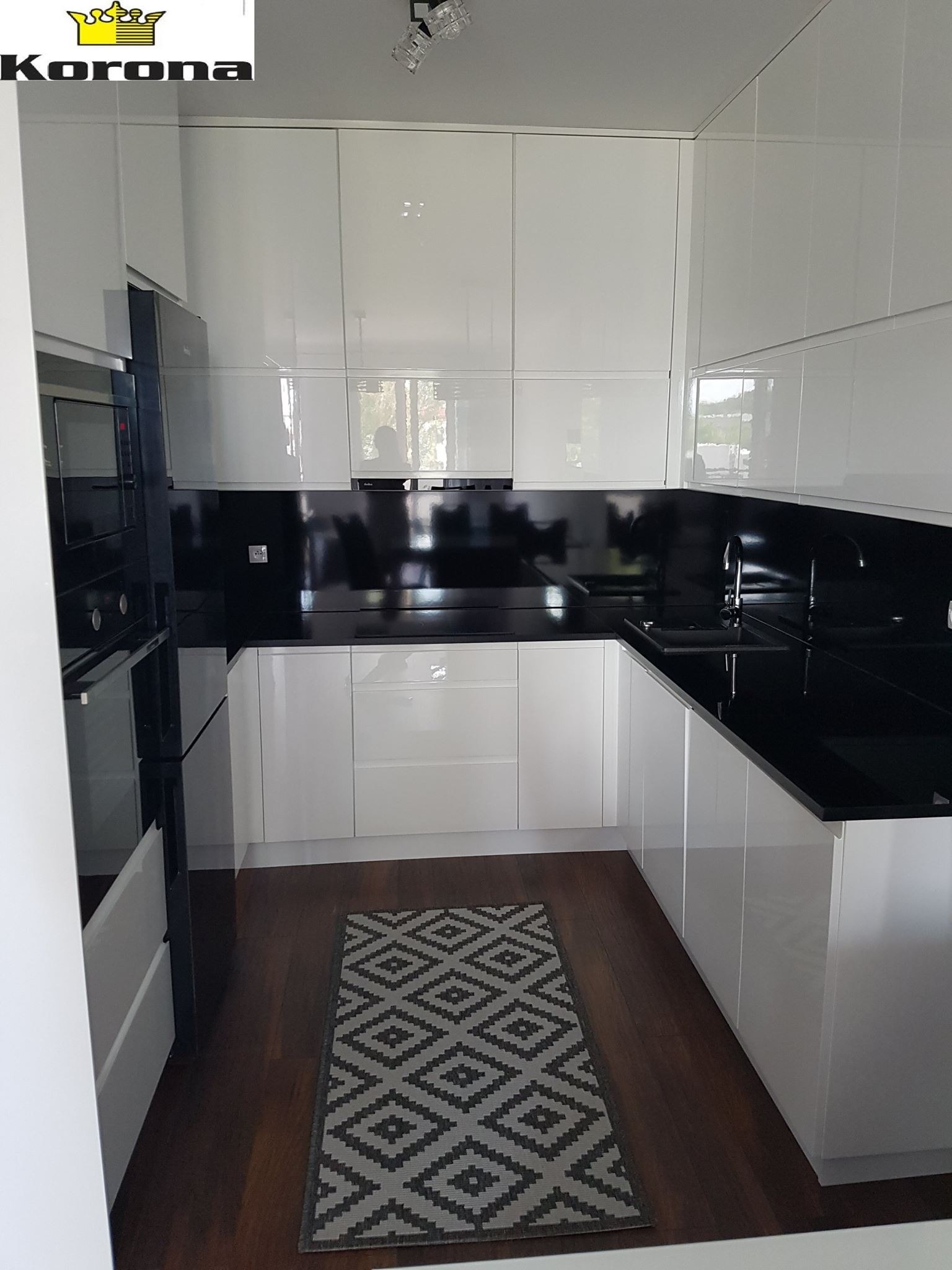 Biała, nowoczesna kuchnia z błyszczącymi frontami szafek, czarnym blatem i panelem nad blatem, oświetleniem punktowym i ciemną podłogą z dywanikiem w geometryczne wzory.
