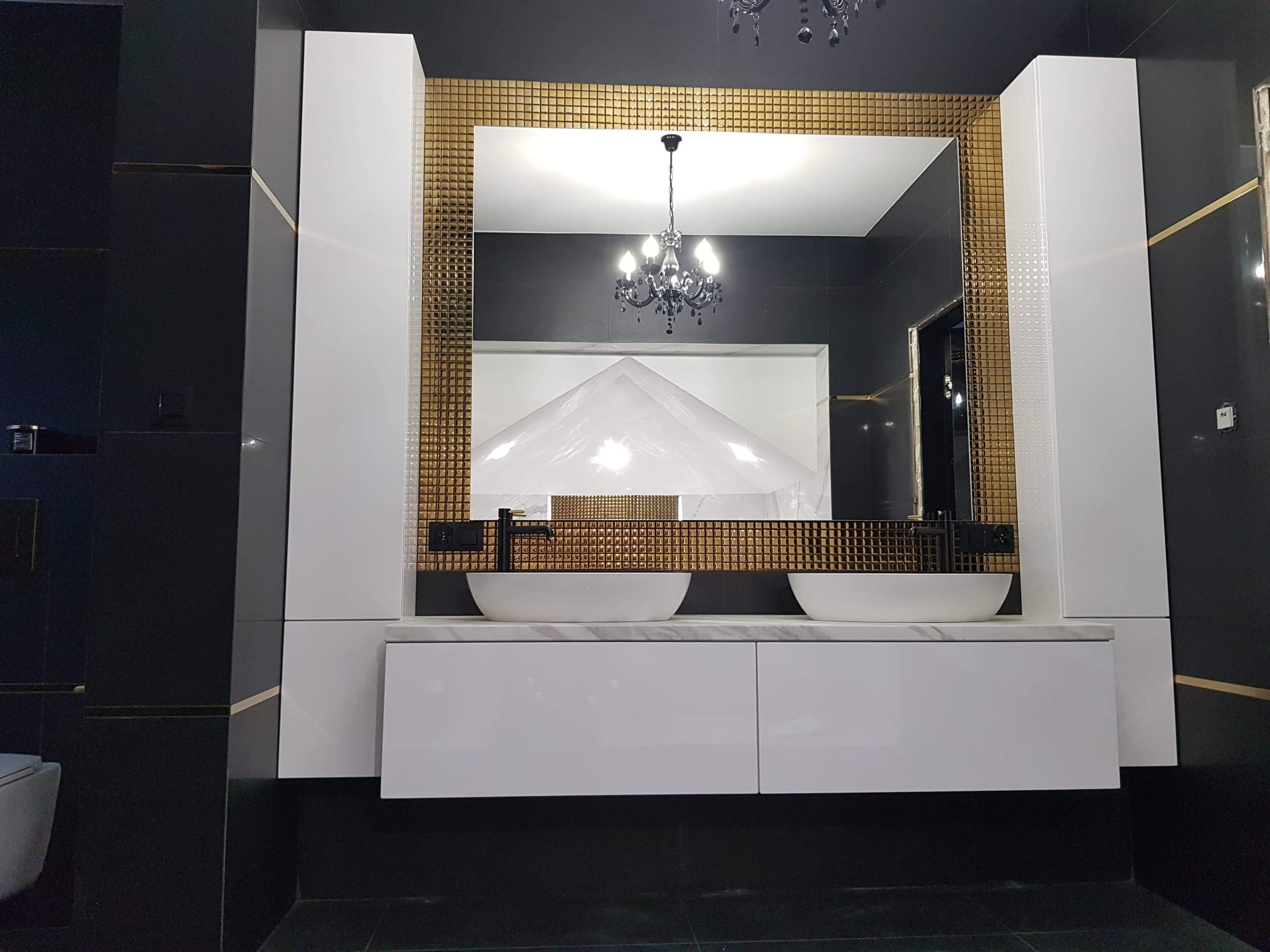 Nowoczesna łazienka z czarnymi ścianami, białymi szafkami pod umywalki i złotą mozaiką wokół lustra, widoczne dwie okrągłe umywalki nablatowe i kryształowy żyrandol odbity w lustrze.