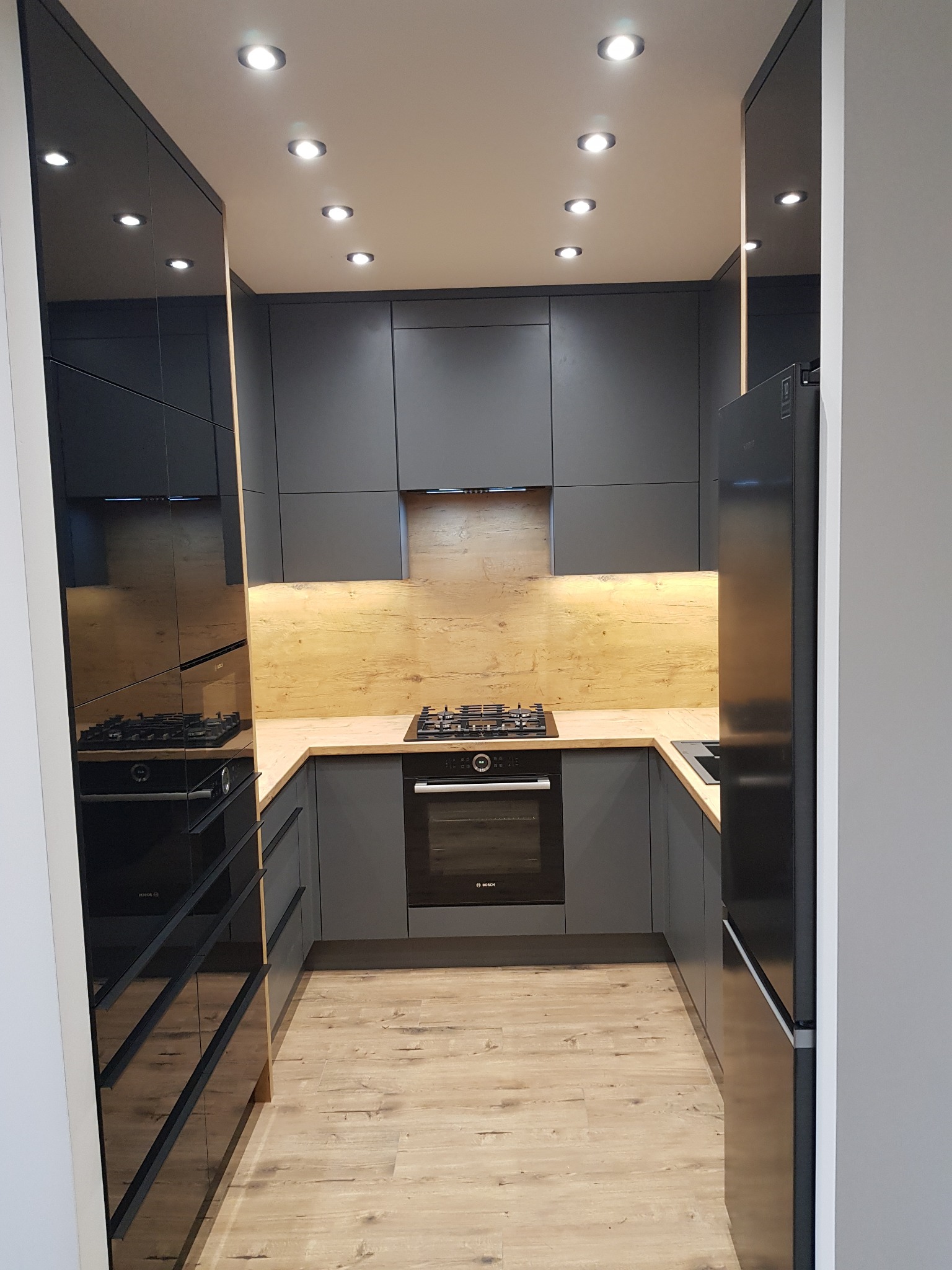 Nowoczesna kuchnia w odcieniach szarości z drewnianymi akcentami, oświetleniem punktowym i błyszczącymi frontami szafek, widziana z perspektywy wejścia.