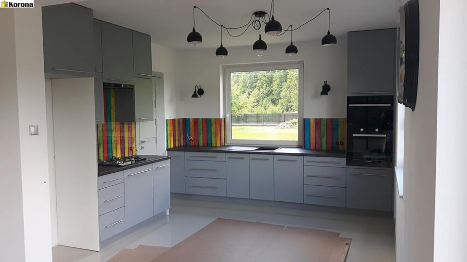 Jasnoszara zabudowa kuchenna z kolorowym panelem nad blatem, widok na okno z zielenią za nim i wisząca lampa z kilkoma czarnymi kloszami.