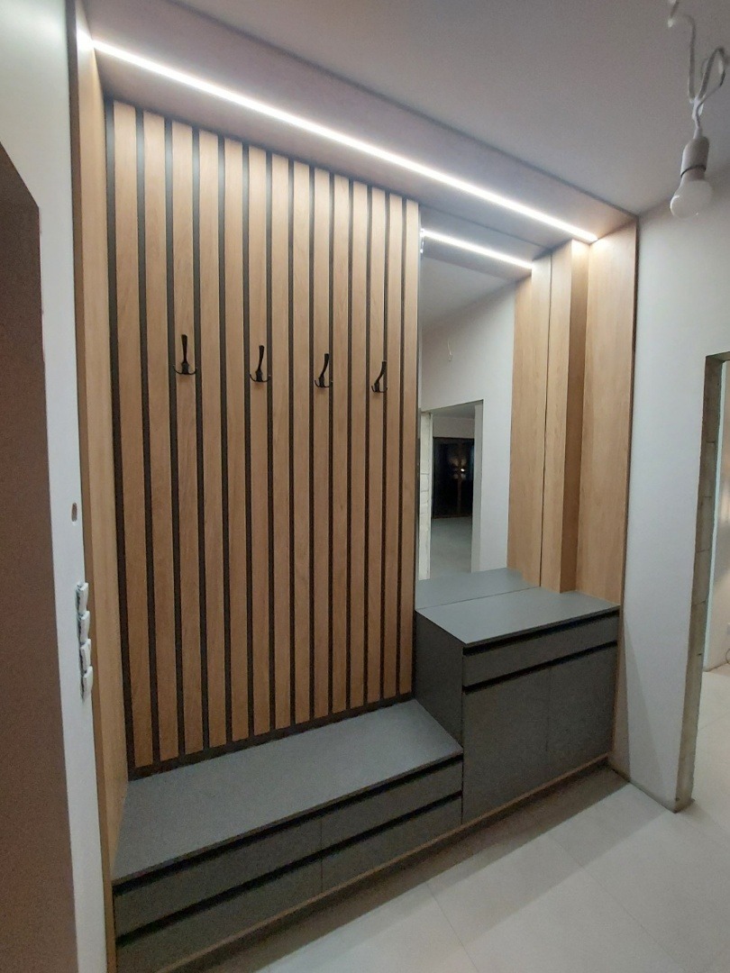 Nowoczesna zabudowa przedpokoju z pionowymi lamelami w odcieniach drewna, wbudowanym siedziskiem i szafką z lustrem, oświetlona listwą LED.