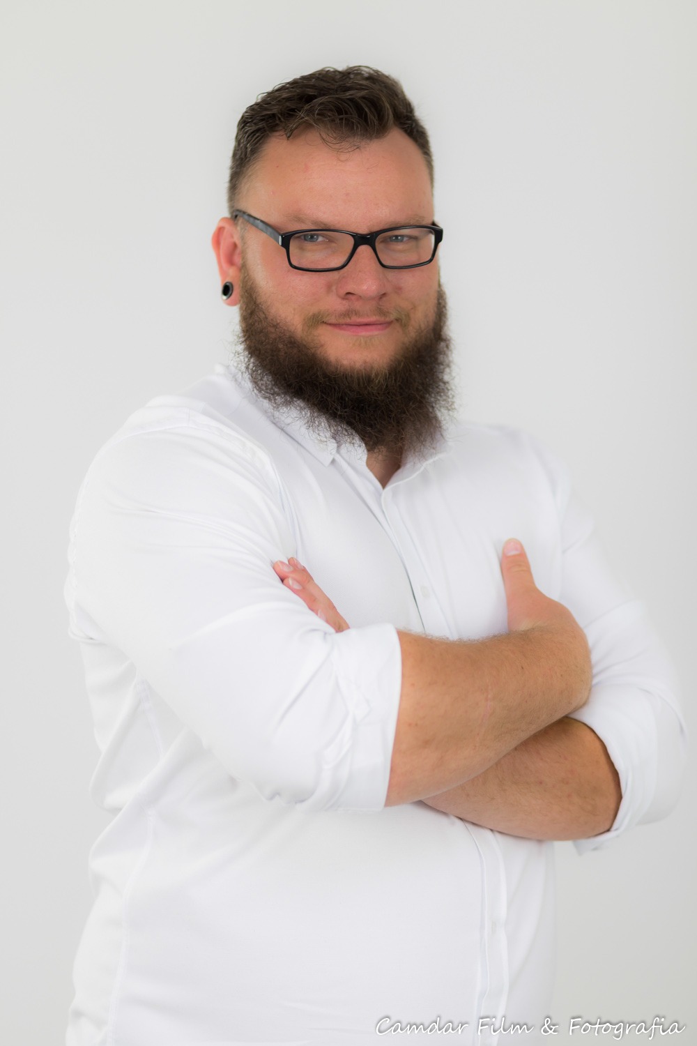 Mężczyzna z brodą i okularami, ubrany w białą koszulę, stoi na białym tle z założonymi rękami.