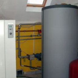 Instalacja grzewcza w kotłowni z żółtą ścianą, widoczne zbiorniki, rury i pompa obiegowa, na ścianie widać ślady wilgoci.