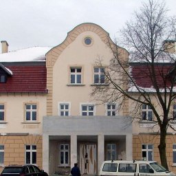 ENERGY PARTNER Bogdan Grala - Duży, dwupiętrowy budynek z czerwonym dachem i beżową fasadą, z kolumnami przed wejściem, częściowo pokryty śniegiem, sfotografowany w pochmurny dzień.