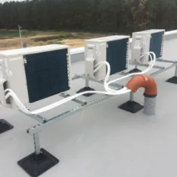 Trzy klimatyzatory na dachu budynku, zamontowane na metalowej konstrukcji. Widoczne białe kable i pomarańczowa rura wentylacyjna. W tle las i zabudowania.