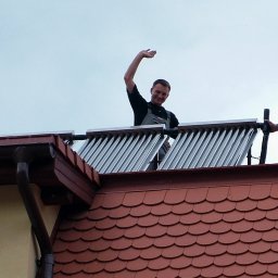 ENERGY PARTNER Bogdan Grala - Montaż kolektorów słonecznych na dachu z czerwoną dachówką. Dwóch uśmiechniętych monterów pozdrawia, jeden w kombinezonie, drugi w stroju roboczym.
