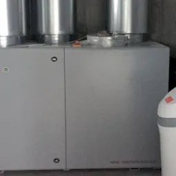 Szara centrala wentylacyjna Salda z dwoma metalowymi przewodami wentylacyjnymi, obok zmiękczacz wody w pomieszczeniu z surowymi ścianami.