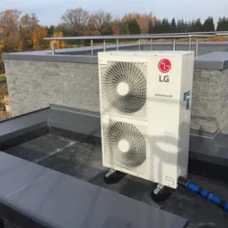 Biała, dwuwentylatorowa jednostka zewnętrzna LG Inverter V na dachu budynku z szarą elewacją i widokiem na jesienne drzewa.