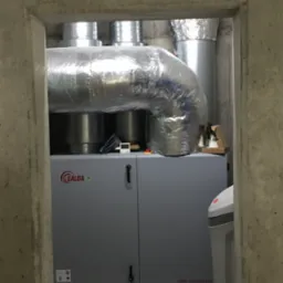 Szara jednostka rekuperacyjna Salda RIRS 1900VER EKO w betonowym pomieszczeniu, z widocznymi srebrnymi kanałami wentylacyjnymi i fragmentem zmiękczacza wody.