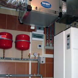 ENERGY PARTNER Bogdan Grala - Złożona instalacja: rekuperator z widocznym logo, zbiorniki wyrównawcze, biała pompa ciepła i rury. Elementy zamontowane na brązowo-beżowej ścianie z płytek.