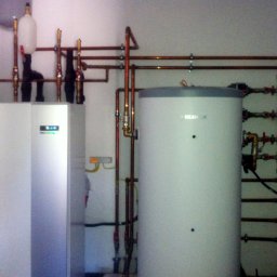 ENERGY PARTNER Bogdan Grala - Biała pompa ciepła z zbiornikiem i miedzianą instalacją w pomieszczeniu. Widoczne liczne rury, zawory i elementy montażowe. Całość sprawia wrażenie nowej instalacji.