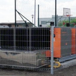 ENERGY PARTNER Bogdan Grala - Kompleksowa instalacja przemysłowej pompy ciepła, zabezpieczona metalowym ogrodzeniem na zewnątrz budynku. Widoczne elementy instalacji i rury doprowadzające.