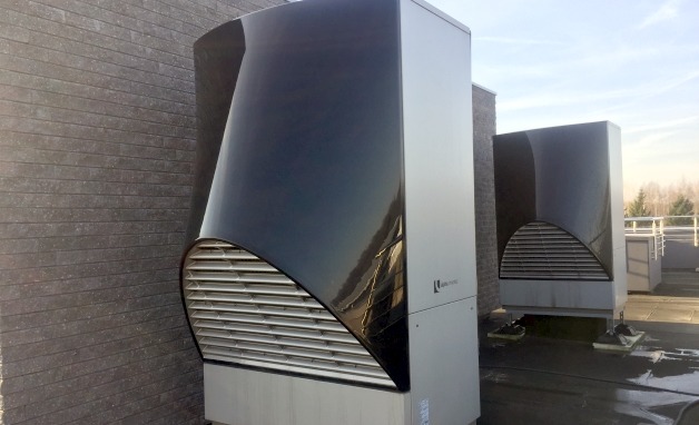 Dwie nowoczesne jednostki zewnętrzne z czarnymi, zaokrąglonymi osłonami wentylatorów, ustawione na dachu budynku z szarą, ceglaną ścianą w tle.