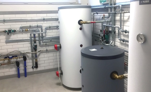 Dwa zbiorniki akumulacyjne, biały i szary, połączone rurami w kotłowni wyłożonej białymi kaflami, z widocznym manometrem i osprzętem hydraulicznym.