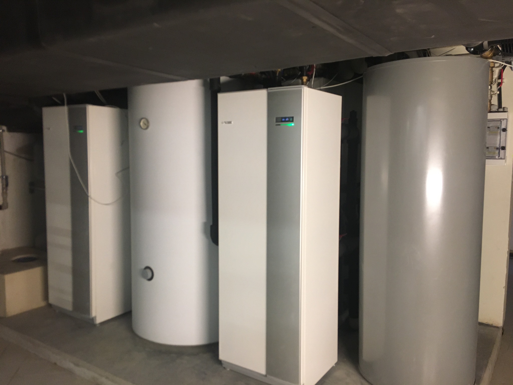 Rząd białych i srebrnych zbiorników, w tym pompa ciepła z zieloną diodą, w pomieszczeniu technicznym z szarą podłogą i niskim sufitem.