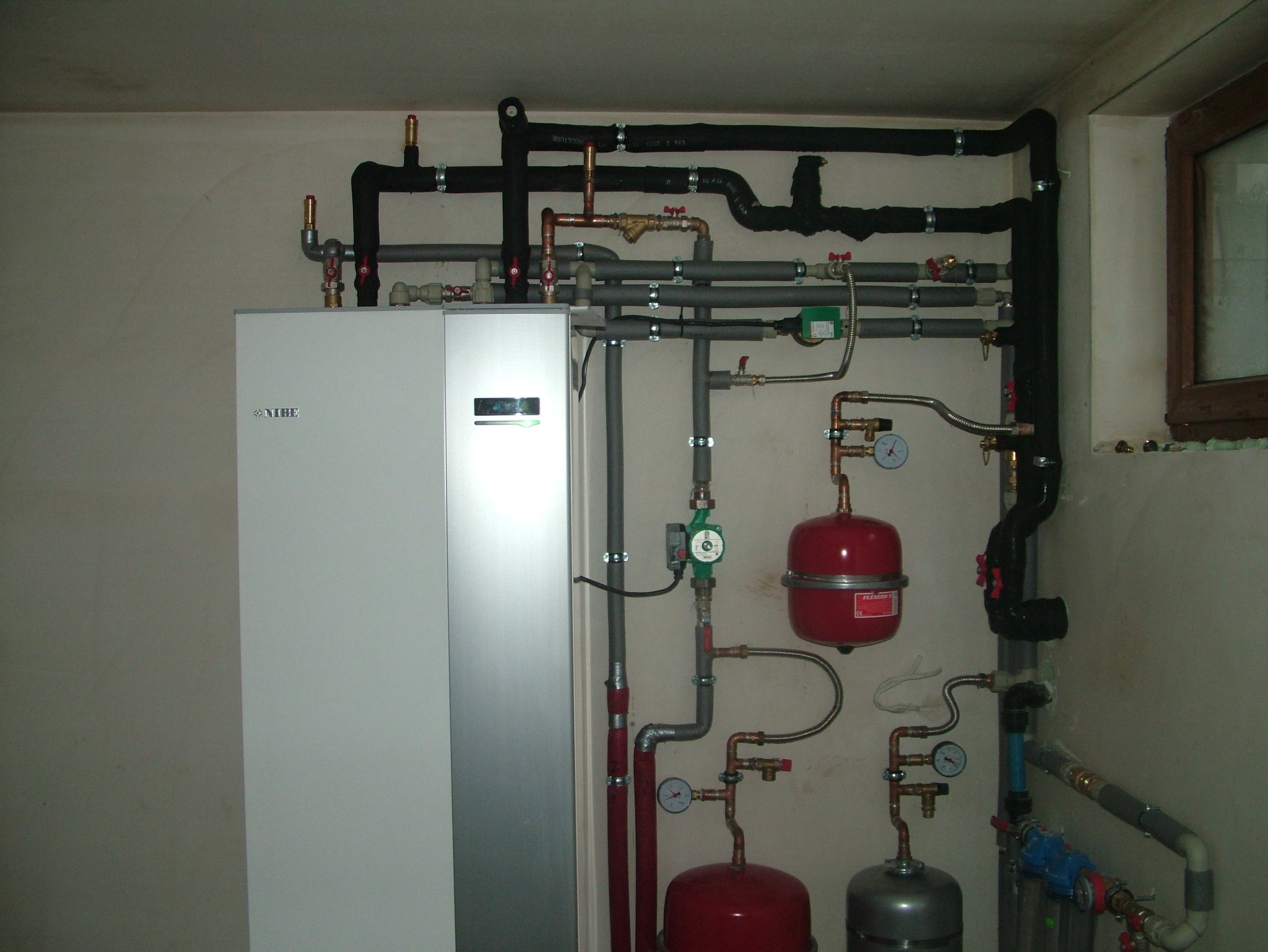 Kompleksowa instalacja grzewcza z szarymi i czarnymi rurami oraz czerwonymi zbiornikami wyrównawczymi, widoczny biały piec grzewczy Nibe i okno.