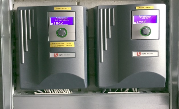 Dwa szare panele sterowania pomp ciepła Alpha Innotec, zamontowane obok siebie, z wyświetlaczami i przyciskami, widoczne okablowanie u dołu.