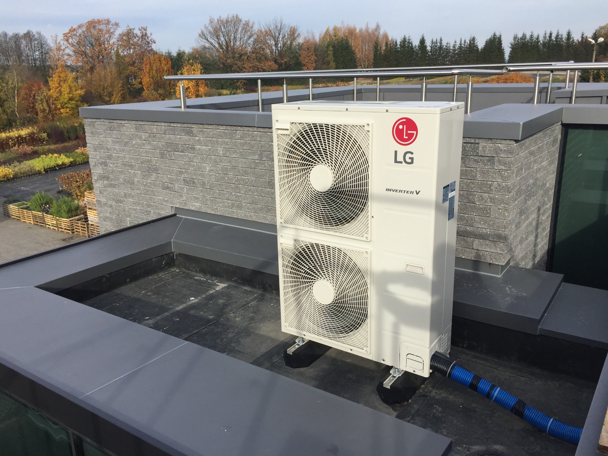 Biała, dwuwentylatorowa jednostka zewnętrzna LG Inverter V na dachu budynku z szarą elewacją i widokiem na jesienne drzewa.