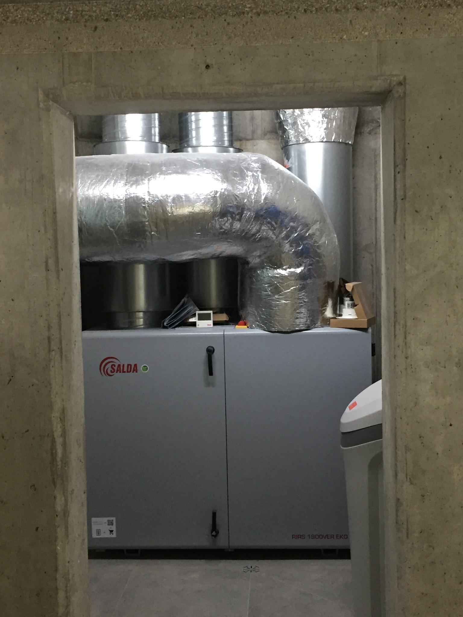 Szara jednostka rekuperacyjna Salda RIRS 1900VER EKO w betonowym pomieszczeniu, z widocznymi srebrnymi kanałami wentylacyjnymi i fragmentem zmiękczacza wody.