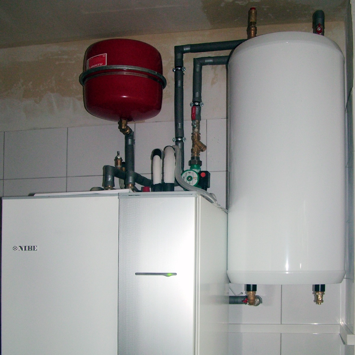 Biały piec NIBE z instalacją: rury, czerwony zbiornik i biały bojler. Ujęcie w kotłowni na tle białych płytek. Widoczny manometr i zawory.