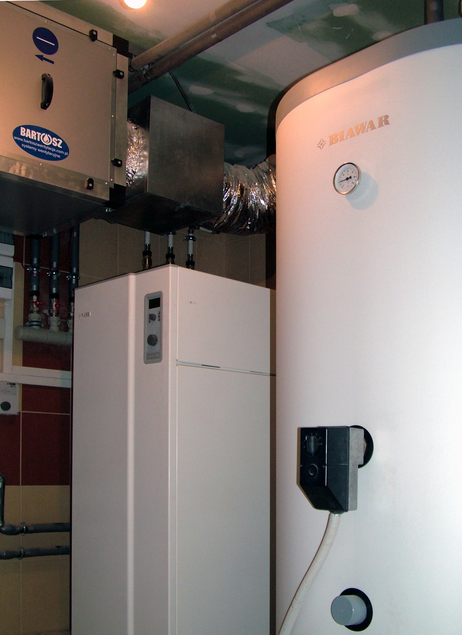 Biały bojler marki Biawar z termometrem, obok rekuperator z logiem Bartosz, widoczne rury i fragment kafelkowanej ściany w tle.