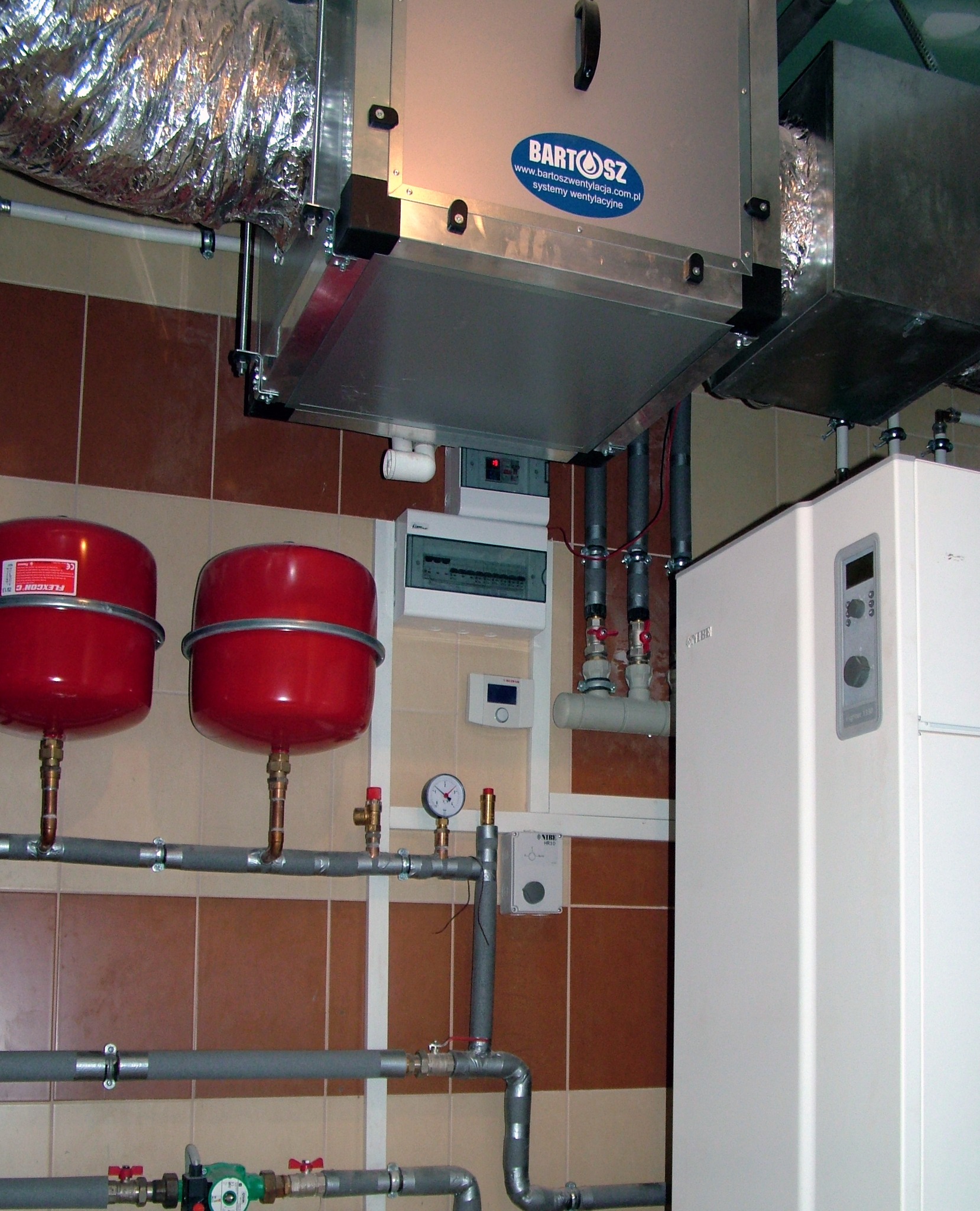 Złożona instalacja: rekuperator z widocznym logo, zbiorniki wyrównawcze, biała pompa ciepła i rury. Elementy zamontowane na brązowo-beżowej ścianie z płytek.